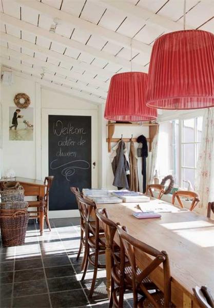 Goście i przyjaciele  spotykają się przy długim drewnianym stole w jadalni. Nadzwyczajnie wyglądają wielkie abażury z cienkiej czerwonej tkaniny.  