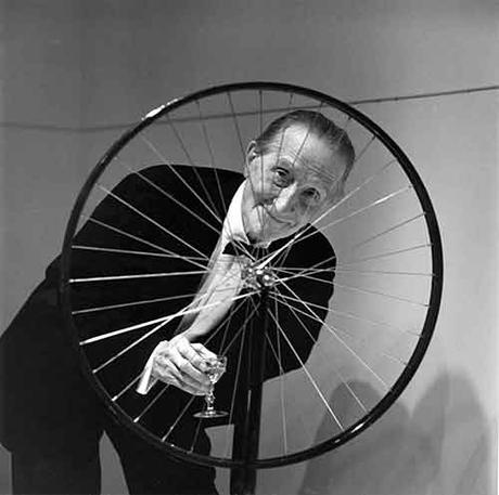Marcel Duchamp sorridente dietro allla sua opera Ruota di bicicletta
